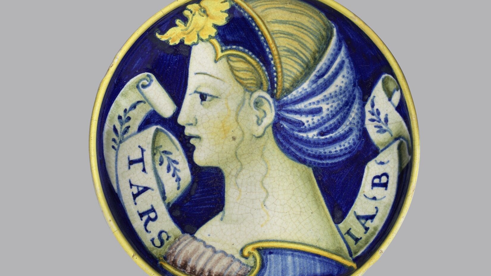 A blue, yellow and white painted dish depicting the profile of a woman with blonde hair and a scroll