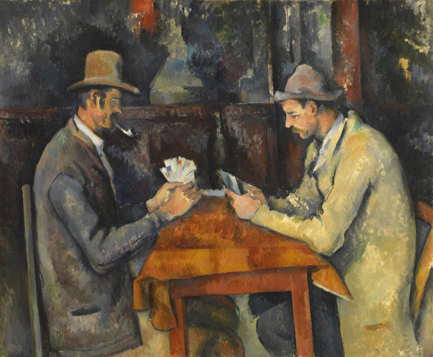 Two men sit across from each other at a table covered with a brown tablecloth, playing cards. Both men wear overcoats and hats, and the man on the left smokes a pipe. They sit inside a wooden building.