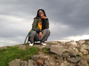 Smiling woman crouching on top of a rocky wall, with dark clouds in the sky behind her.