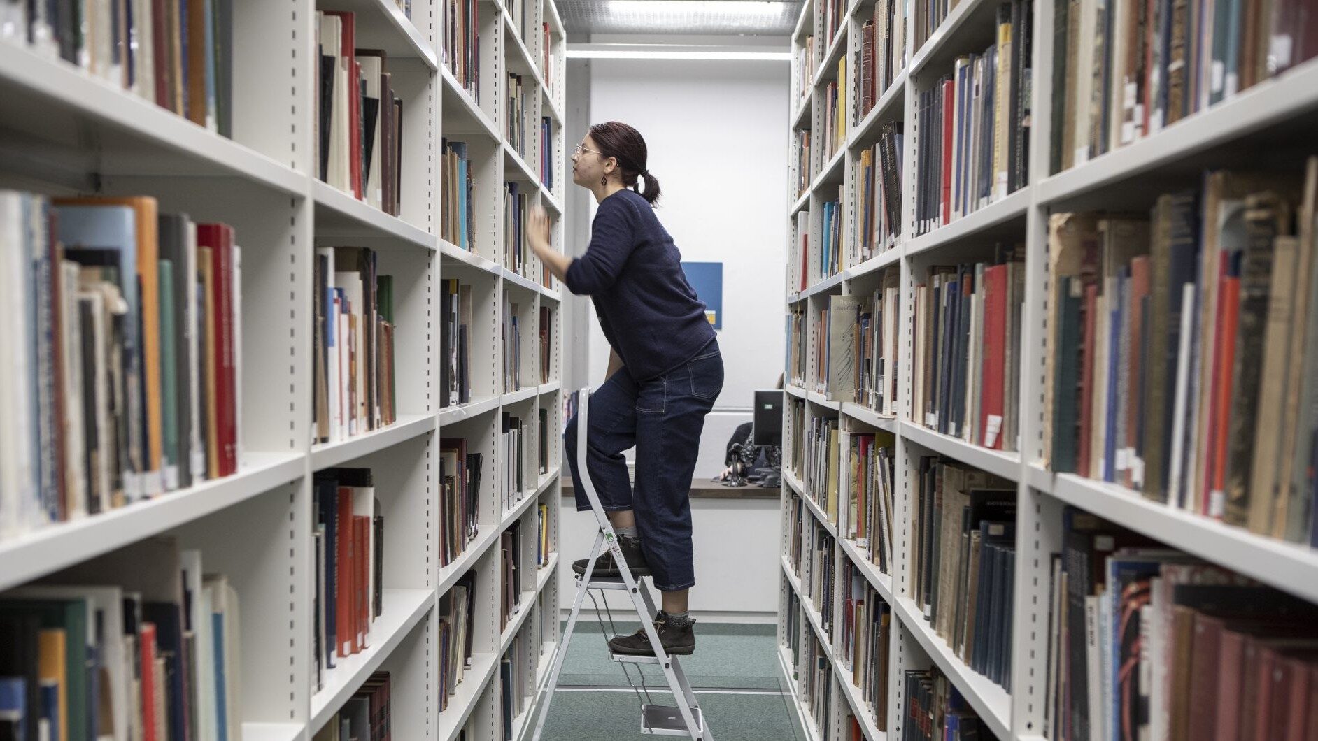 student climbing a step ladder to reach higher shelves of books in the library