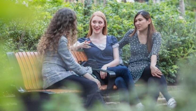 Three students sitting on a bench, chatting, surrounded by greenery, at The Courtauld's Vernon Square campus.