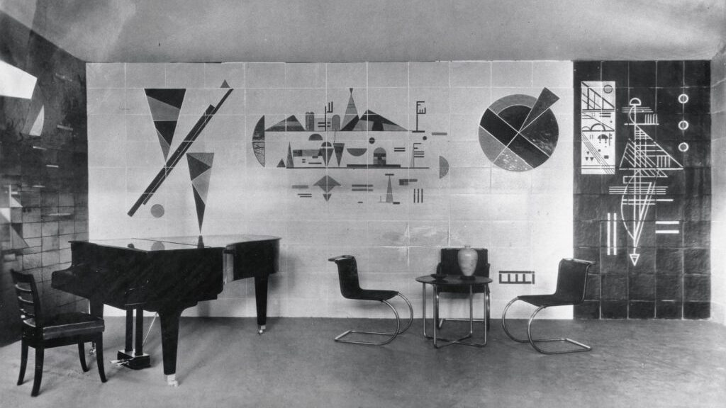 photograph of a room with three chairs and a piano, there are geometric drawings and prints on the wall