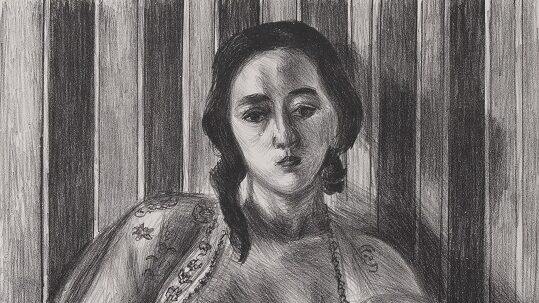 A nude woman sits on the floor, left leg drawn to her chest and right leg outstretched, with a vacant gaze. She is wearing a sheer, embroidered, open blouse. The wall behind her has a dark and light vertical stripes, and on the floor she sits on a flowery embroidered rug. A signature indicates the print is 33/50.