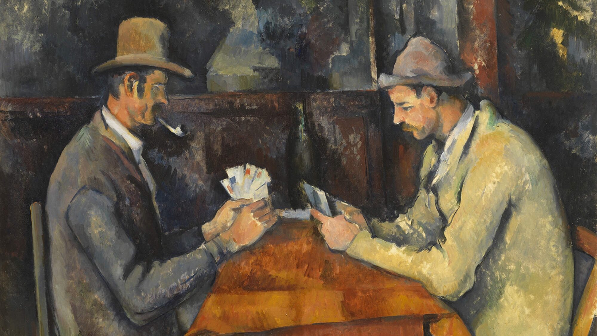 Two men sit across from each other at a table covered with a brown tablecloth, playing cards. Both men wear overcoats and hats, and the man on the left smokes a pipe. They sit inside a wooden building.