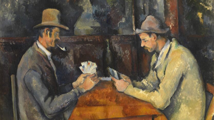 Two men sit across from each other at a table covered with a brown tablecloth, playing cards. Both men wear overcoats and hats, and the man on the left smokes a pipe. They sit inside a wooden building.