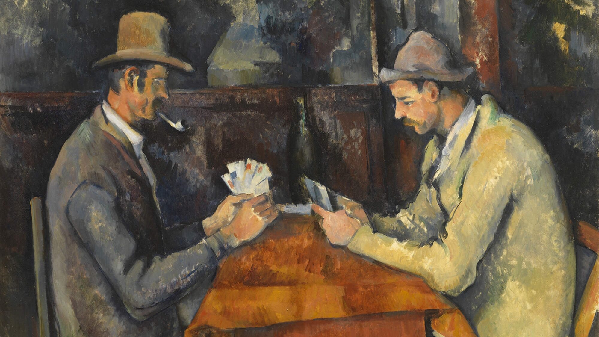 Two men sit across from each other at a table covered with a brown tablecloth, playing cards. Both men wear overcoats and hats, and the man on the left smokes a pipe. They sit inside a wooden building.