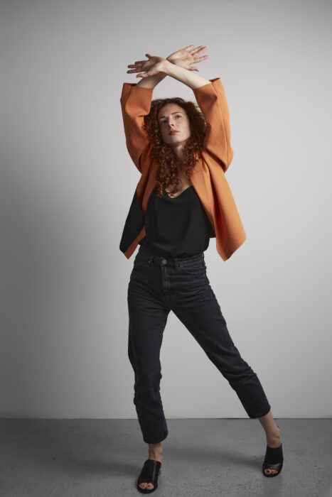 Photograph of a woman in an orange jacket with her arms raised above her head