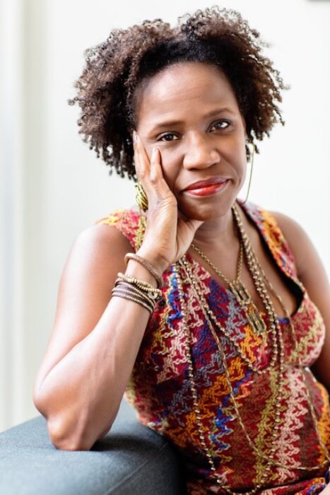 Photograph of a black woman facing the camera, leaning on a wall with her elbow, hand on cheek