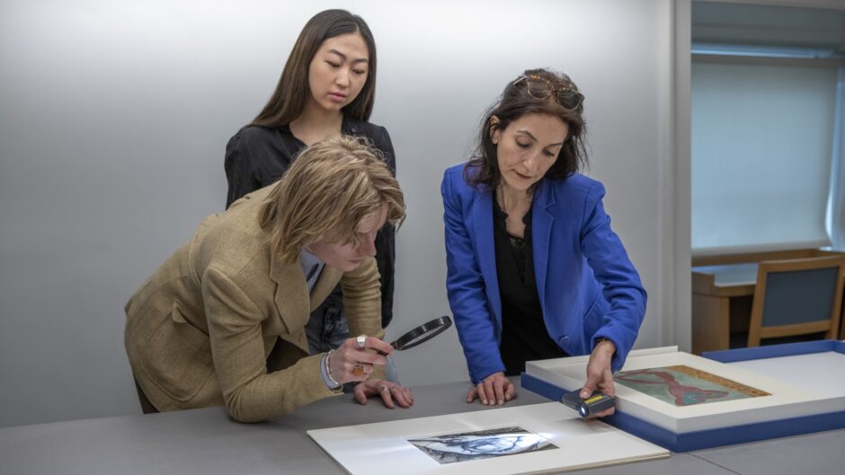 Three people examining artwork on a table with magnifying tools.