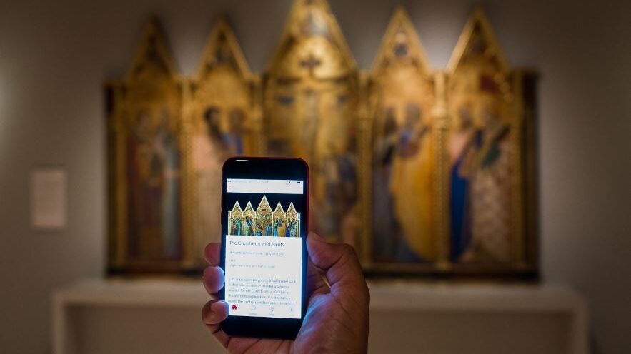 An image of a visitor holding up their phone with the Bloomberg Connects guide showing, in the gallery