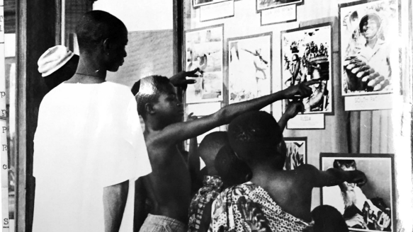 Photograph showing a group of Black children and adults looking into a window and pointing. Behind the window are various photographs on display