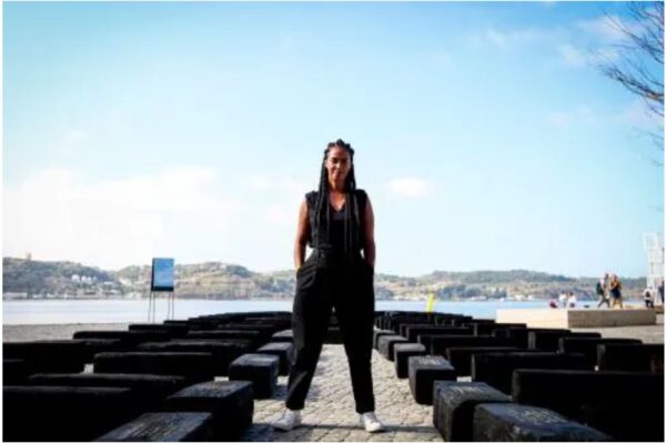 Photograph showing a black woman with long, braided hair standing against a blue sky and among black blocks of wood
