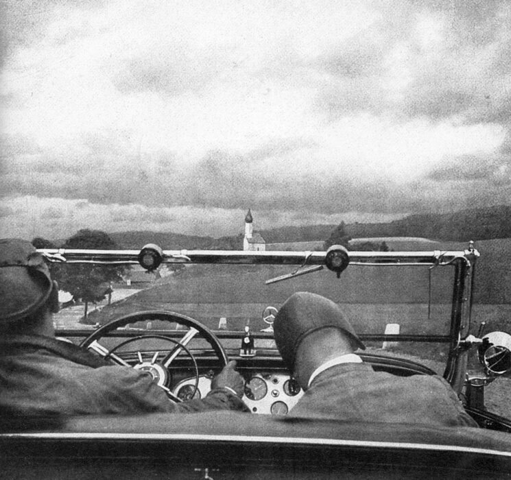 Photograph showing the backs of the heads of two male figures sitting in the front two seats of an open top car