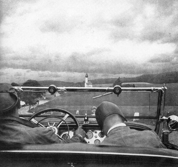 Photograph showing the backs of the heads of two male figures sitting in the front two seats of an open top car