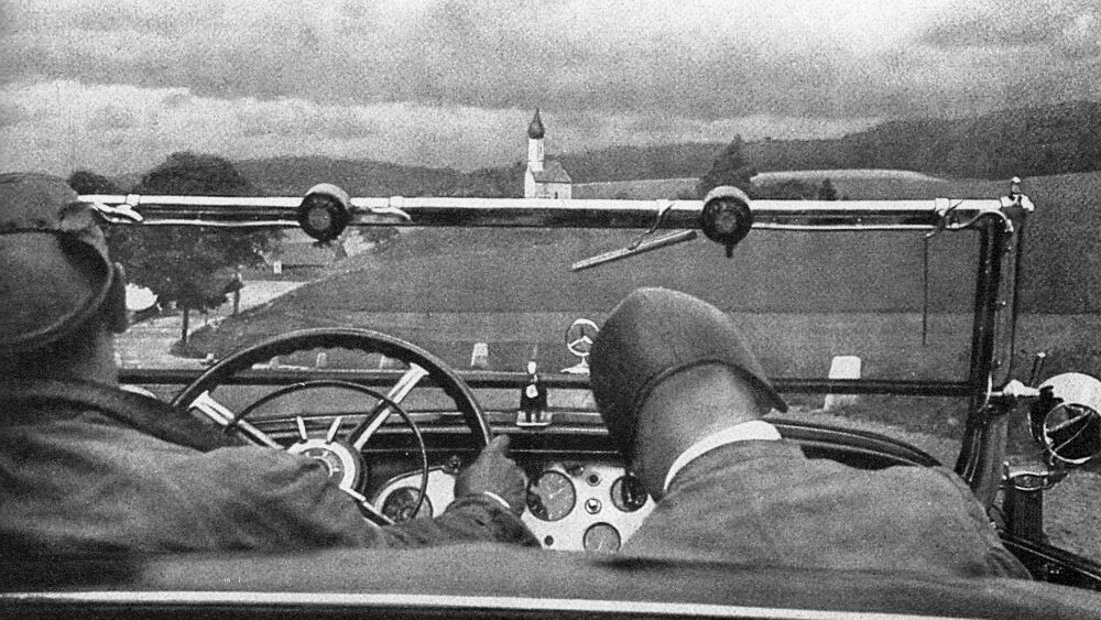 Photograph showing the backs of the heads of two male figures sitting in the front two seats of an open top car