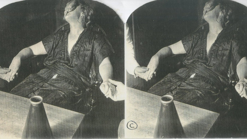 A stereoscopic image showing a dual image of a woman sat at a table with her head back, face covered by her wet hair, while she holds hands with figures on either side of her