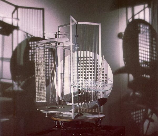 Photograph of a sculpture made from metal shapes and frames. Lights are shining in different directions on it, casting large geometric shadows on the wall behind.