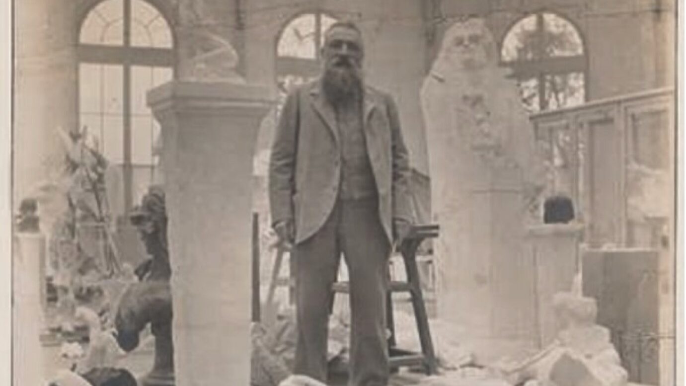 Black and white photograph of the artist Rodin standing on a platform in his studio surrounded by parts of sculptures