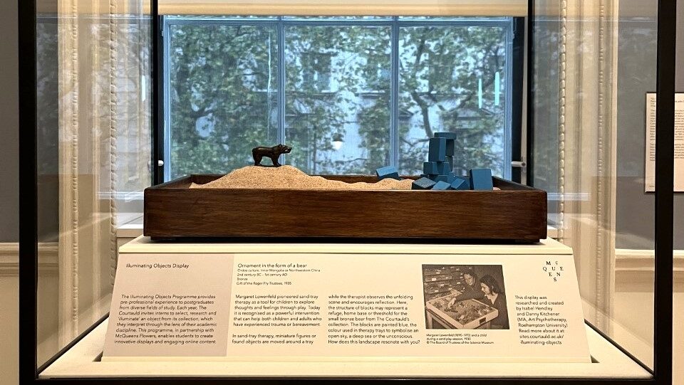 Photograph of a gallery display showing a sandbox with a carved bear.