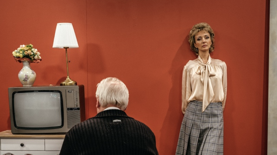 Woman standing against a red wall as man in front of her stares at a TV