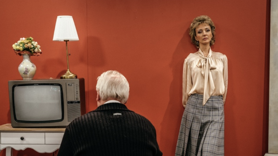 Woman standing against a red wall as man in front of her stares at a TV