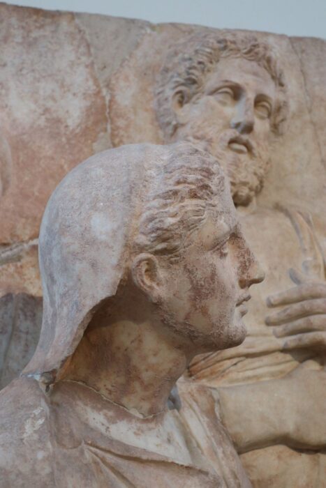 A sculpture of a woman whose head is turned to the right. In the background is a sculpture of a man clutching his stomach.