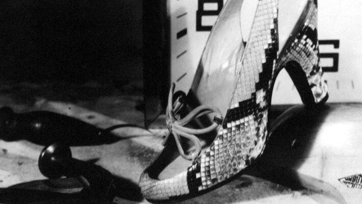 Photograph showing a snakeskin high heeled shoe in front of a clock