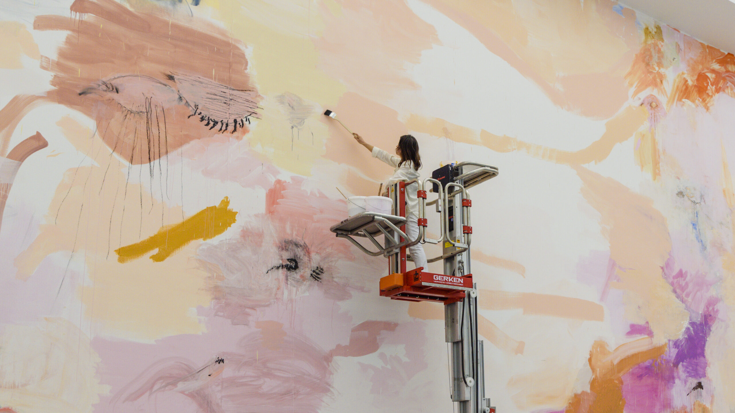 Photograph showing a woman on top of a mechanical raised platform, adding paint to a painting which fills the entire wall of the room