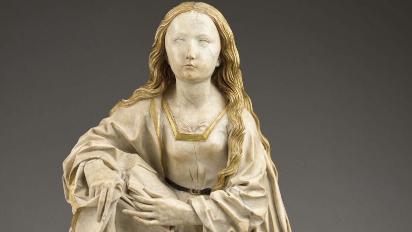 Photograph of a sculpture of the Virgin Mary, who leans on a book sitting on a plinth