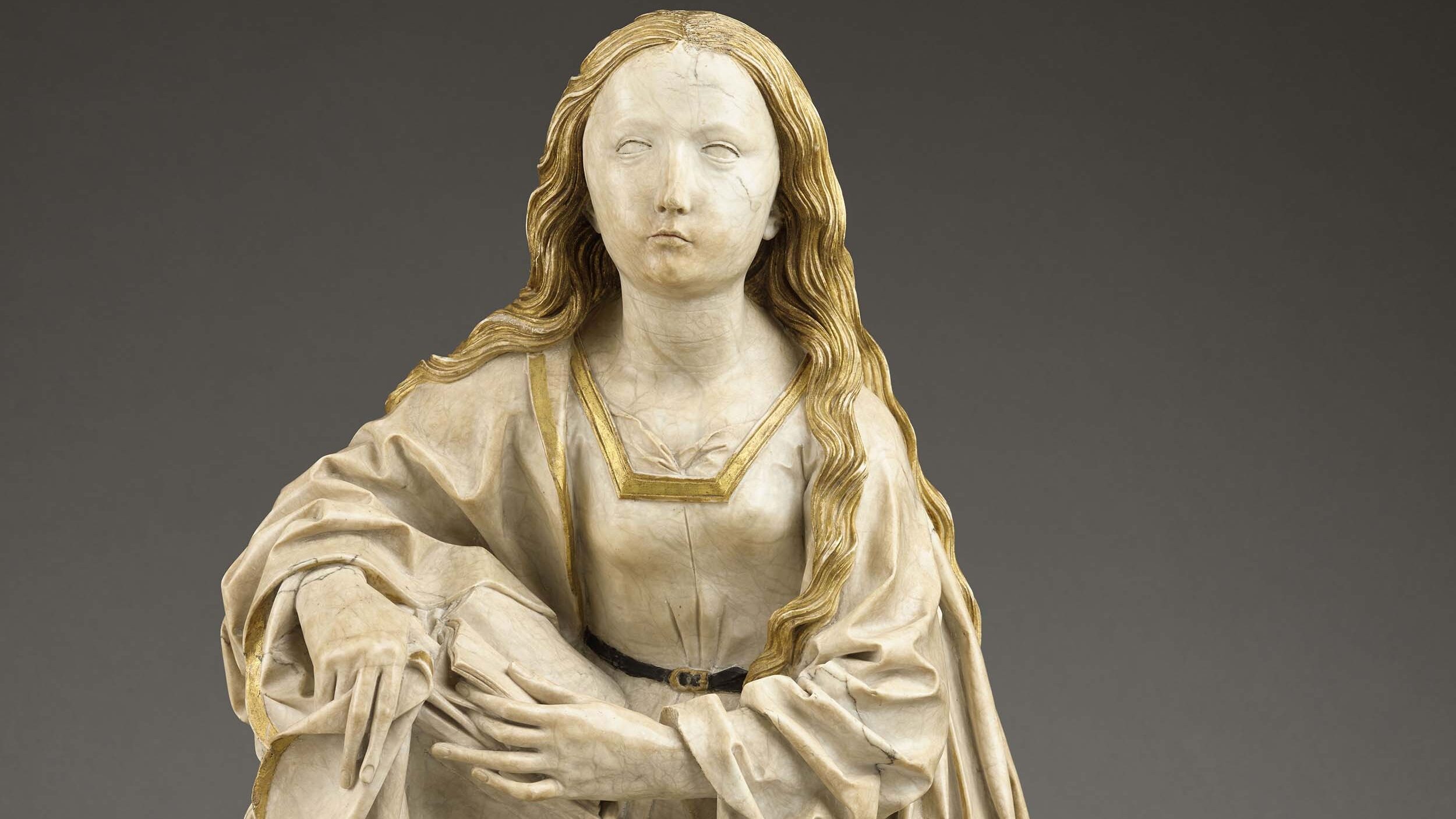 Photograph of a sculpture of the Virgin Mary, who leans on a book sitting on a plinth
