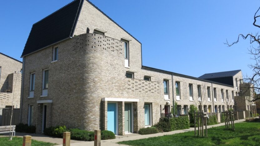 Photograph of a grey building against a blue sky. There are bollards to the left of the building and grass and trees in front of it.