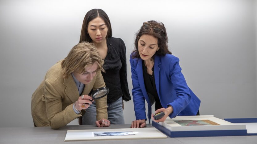 group of three students looking at a print on a table using a magnifying glass