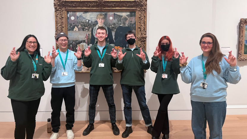 a group of six gallery assistant standing in front of one of the painting in the Courtauld's gallery smiling and crossing their fingers