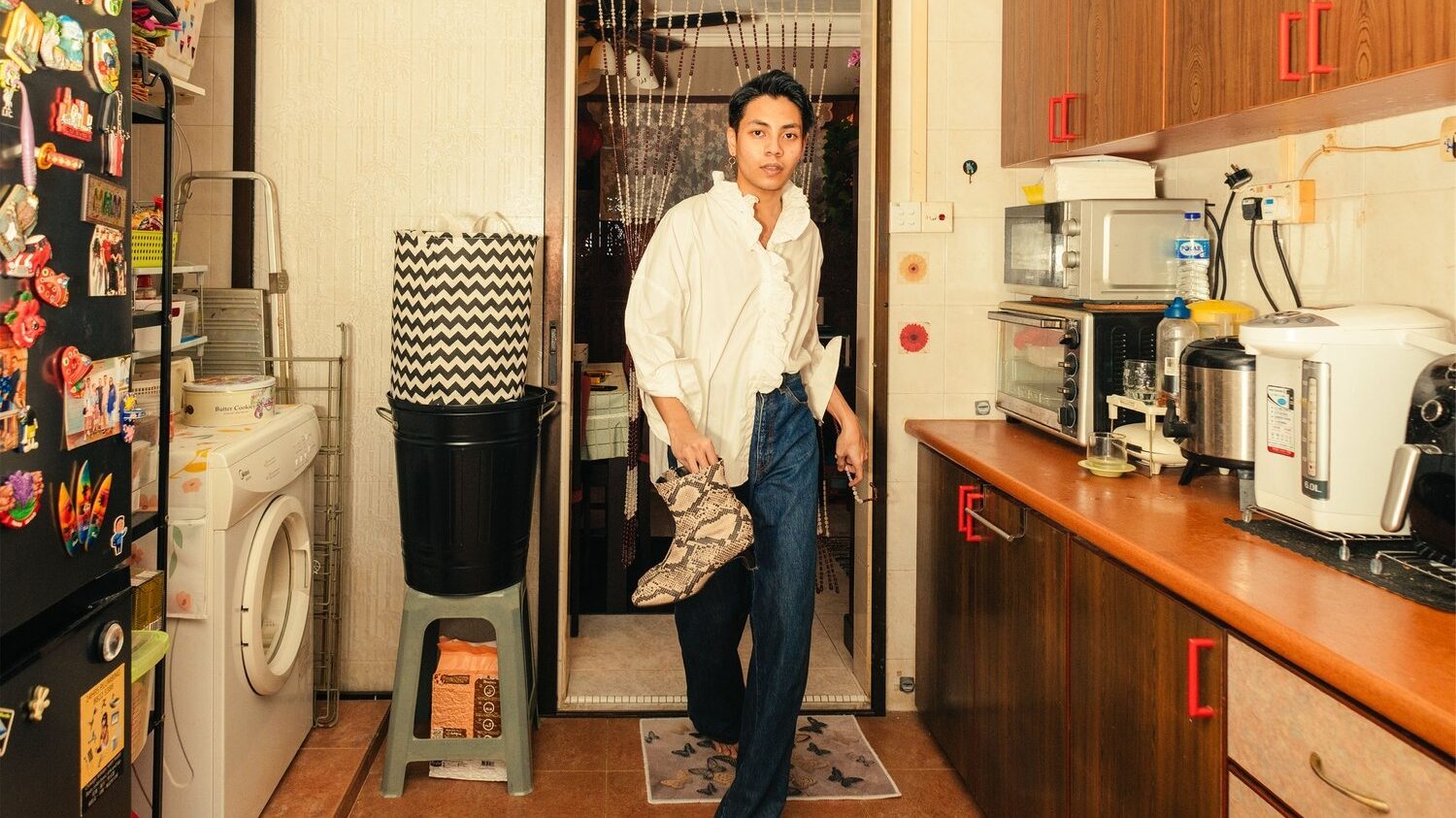 A dark haired man walks towards the camera through a beaded curtain. He is walking into a kitchen, barefoot, and is carrying a pair of snakeskin boots with him.