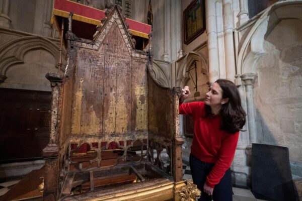 Krista, with dark brown hair and wearing a red jumper, carries out conservation work on the Coronation Chair, shown in full and up on a stand.