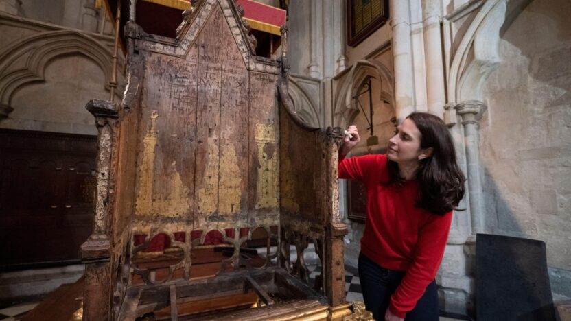 Krista, with dark brown hair and wearing a red jumper, carries out conservation work on the Coronation Chair, shown in full and up on a stand.