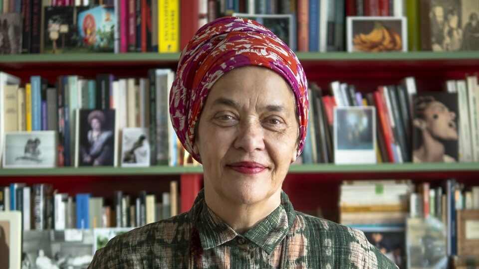 Photograph of a black woman standing with her hands on her hips against a backdrop of a bookcase.