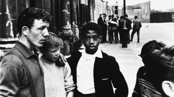Two groups of men and boys on Southam Street, North Kensington, London. A young West Indian boy looks straight at the camera.