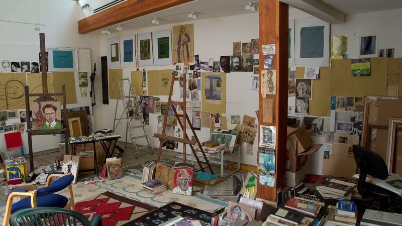 Photograph of the interior of an artist's studio. The walls and floor are covered with paintings, drawings, and books and there are two easels central.