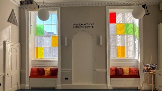 Still of the Leon Kossoff Learning Centre with no tables or chairs. Light streams in from two large windows in the ornate but friendly room; the windows have three and four scattered panes respectively (out of twelve total on each window) that have brightly coloured glass in them.