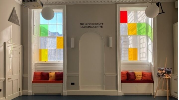 Still of the Leon Kossoff Learning Centre with no tables or chairs. Light streams in from two large windows in the ornate but friendly room; the windows have three and four scattered panes respectively (out of twelve total on each window) that have brightly coloured glass in them.