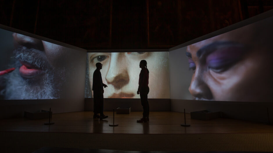 Photograph showing an installation of three video screens with two people in sillhouette against them
