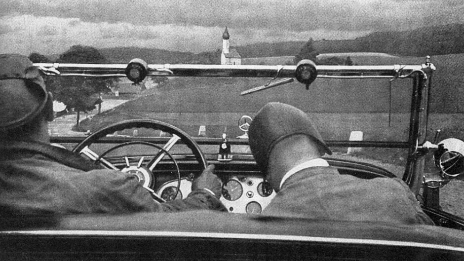 Photograph showing the backs of the heads of two male figures sitting in the front two seats of an open top car