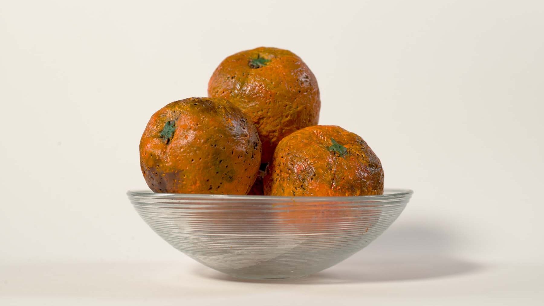 Four handmade, clay oranges are placed in a bowl. White background.