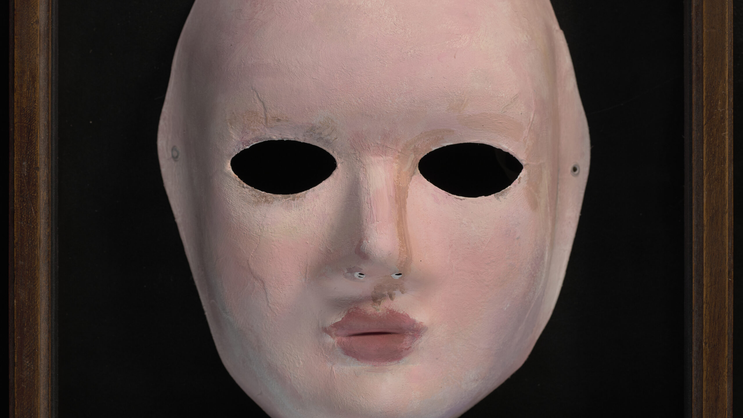 A paper mache mask. Pink in colour, photographed on a black background.