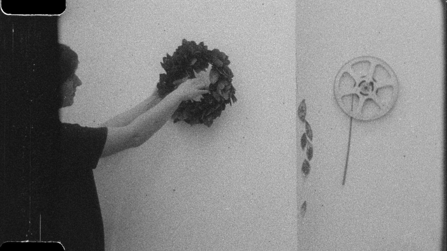 A still from the film 'Daphne' showing a woman holding a laurel wreath. She stands against a white wall.