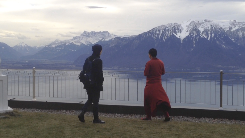 two people standing in front of a fence overlooking a lake and some mountains