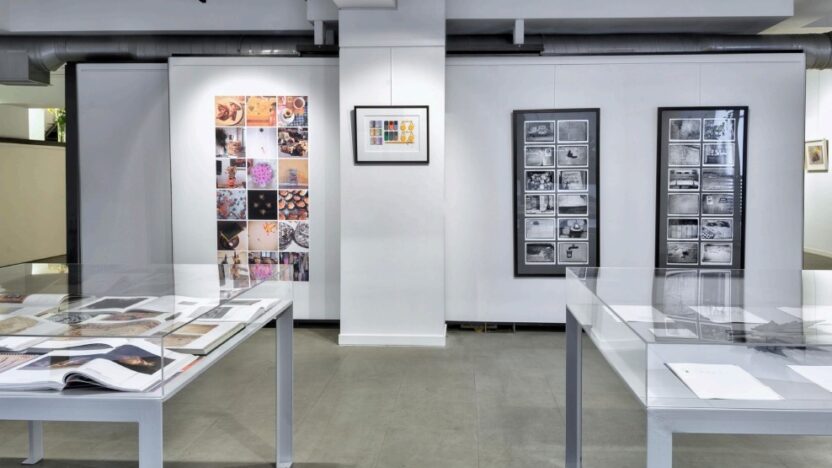 Exhibition view of the Sotiris Felios Collection space, with white walls, collage artworks on the back wall, and two display tables visible in the foreground.