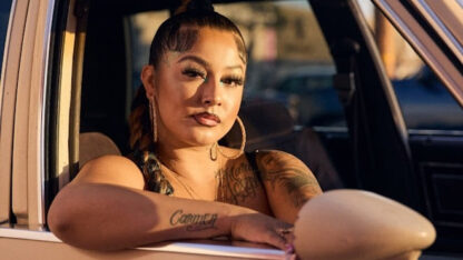 Photograph of a woman sitting in a peach-coloured vintage car, looking at the camera, bathed in evening sunlight. She has dark hair tied back in a braid, large gold earrings, and tattoos on her arms.