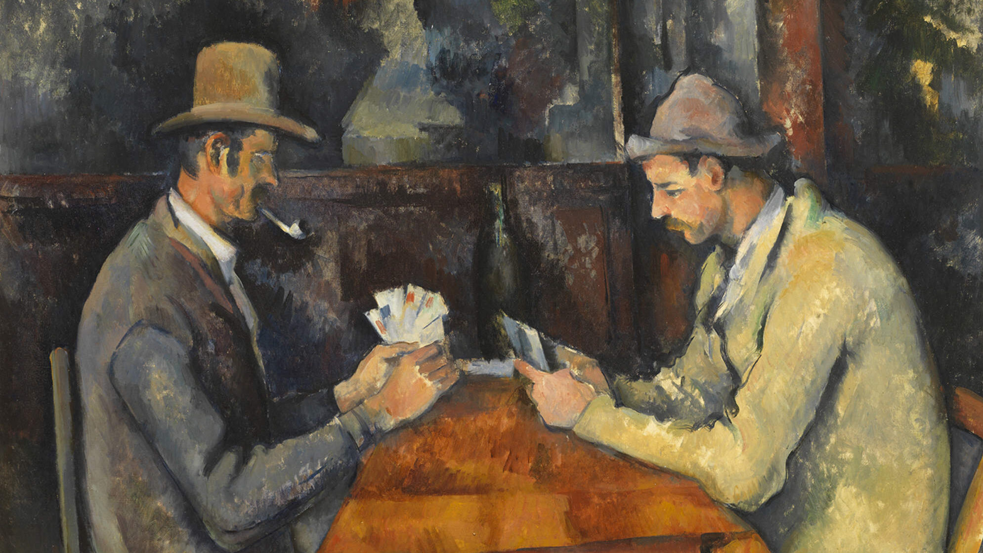 Two men sit across from each other at a table covered with a brown tablecloth, playing cards. Both men wear overcoats and hats, and the man on the left smokes a pipe. They sit inside a wooden building.